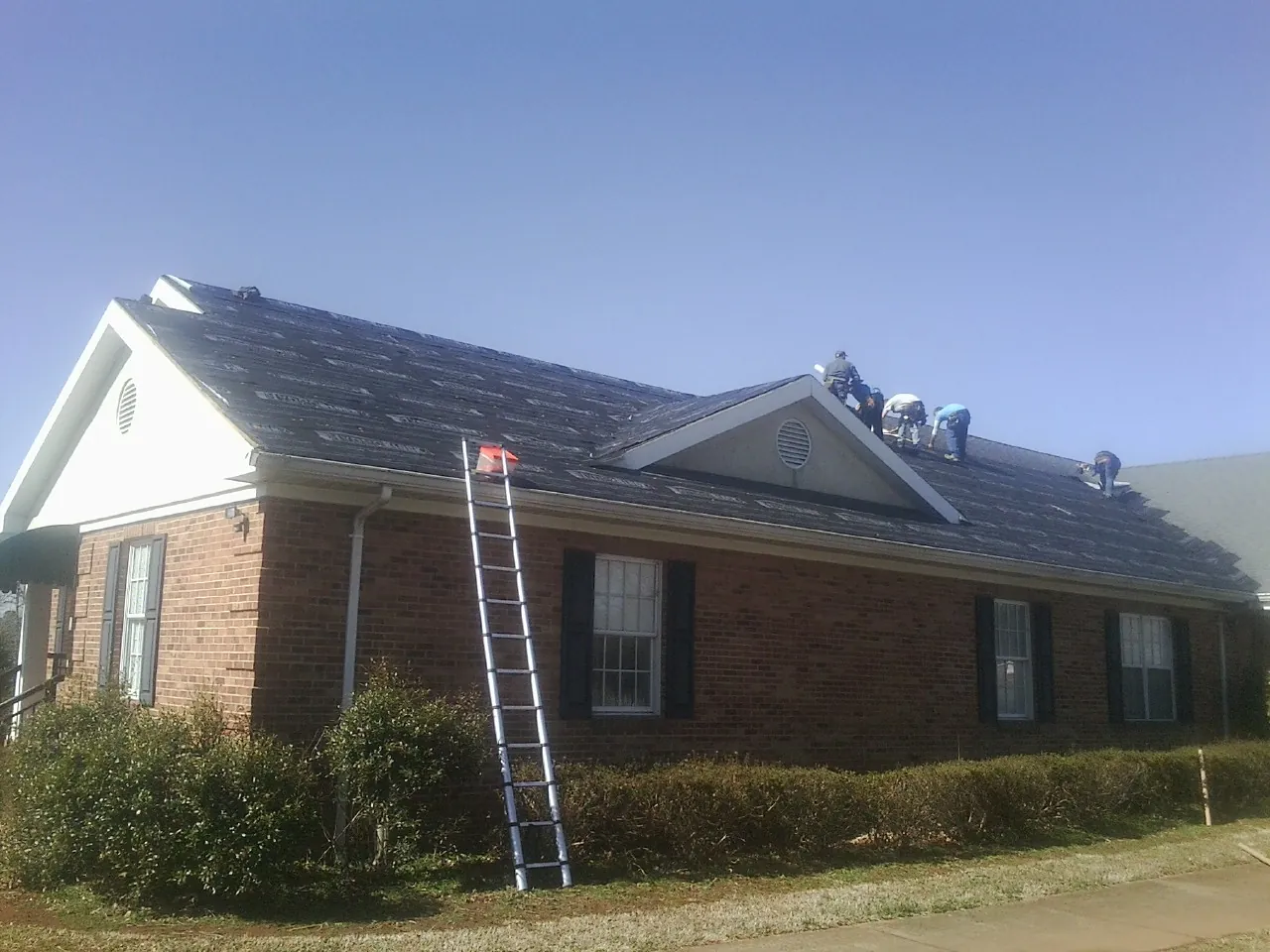 Crew tearing shingles off a brick ranch roof with a ladder set — Metal Roof Repair work by our crew in Blossvale