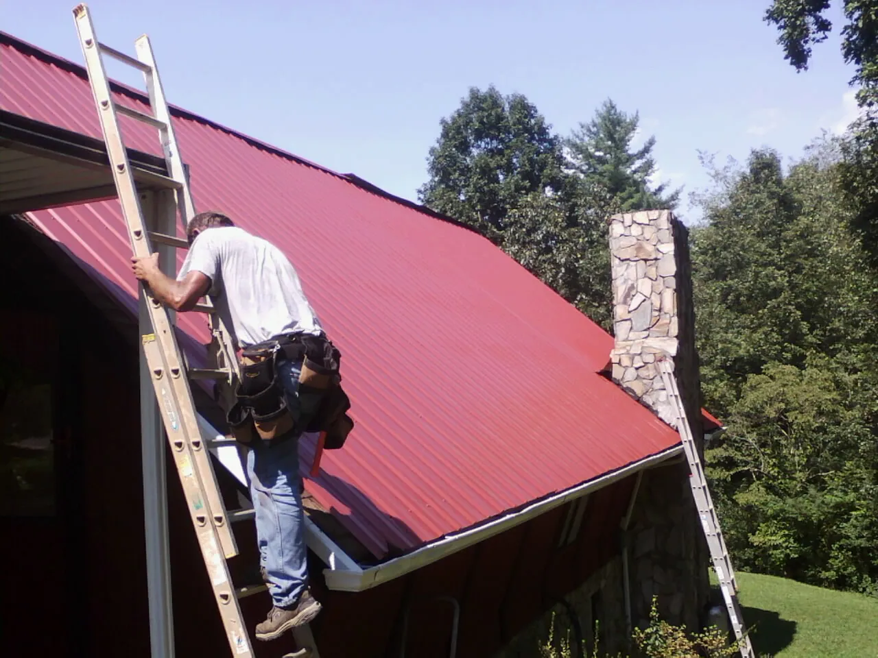 Roofer on a ladder beside a red metal roof and stone chimney — Soffit & Fascia Repair by Roofing Brothers in Blossvale
