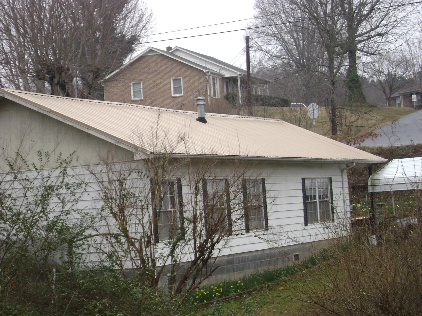 Tan metal roof on a white sided house set into a hillside — Commercial Roofing by Roofing Brothers in Blossvale